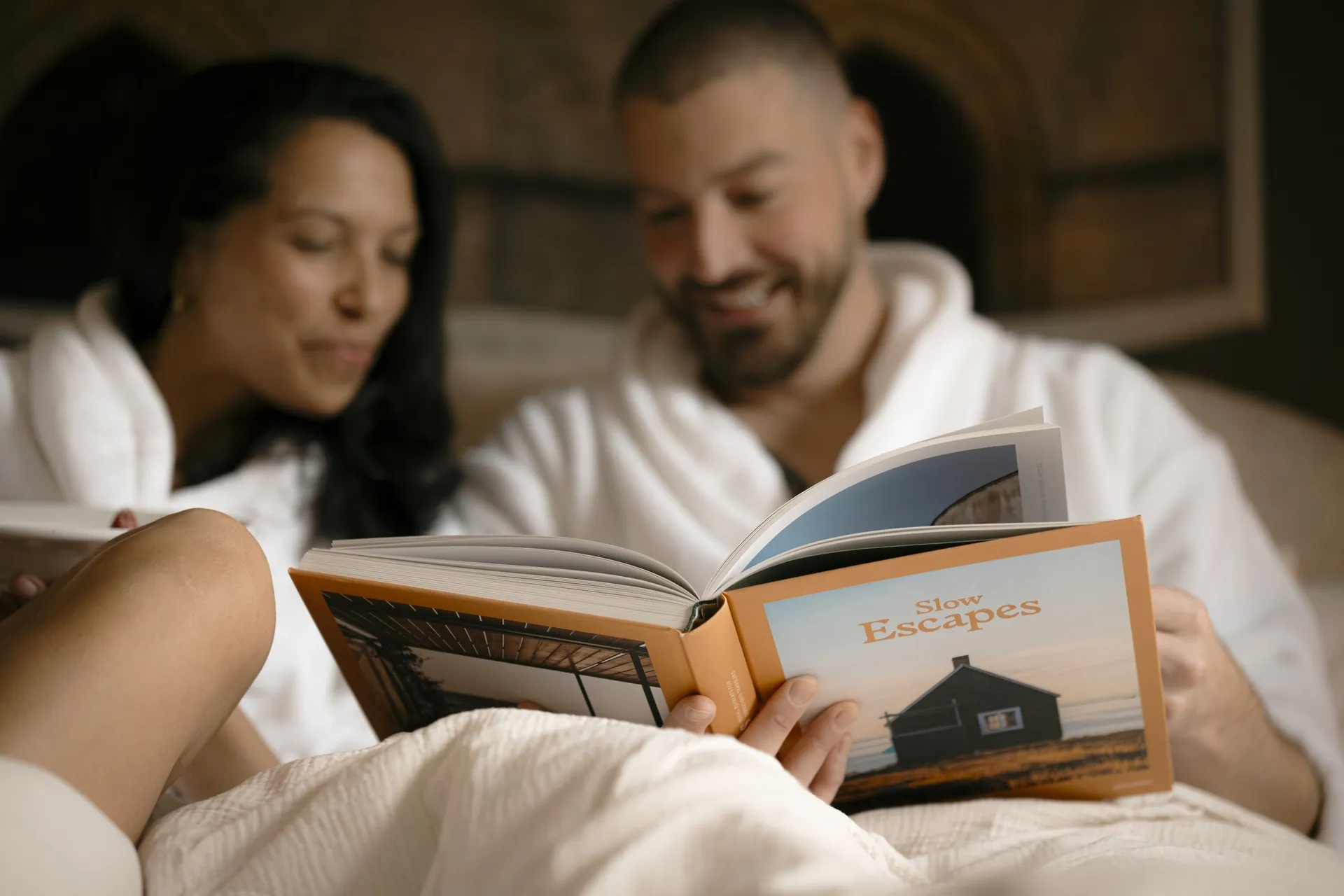 Two guests in white robes reading a Slow Escapes book together in bed at Ralia