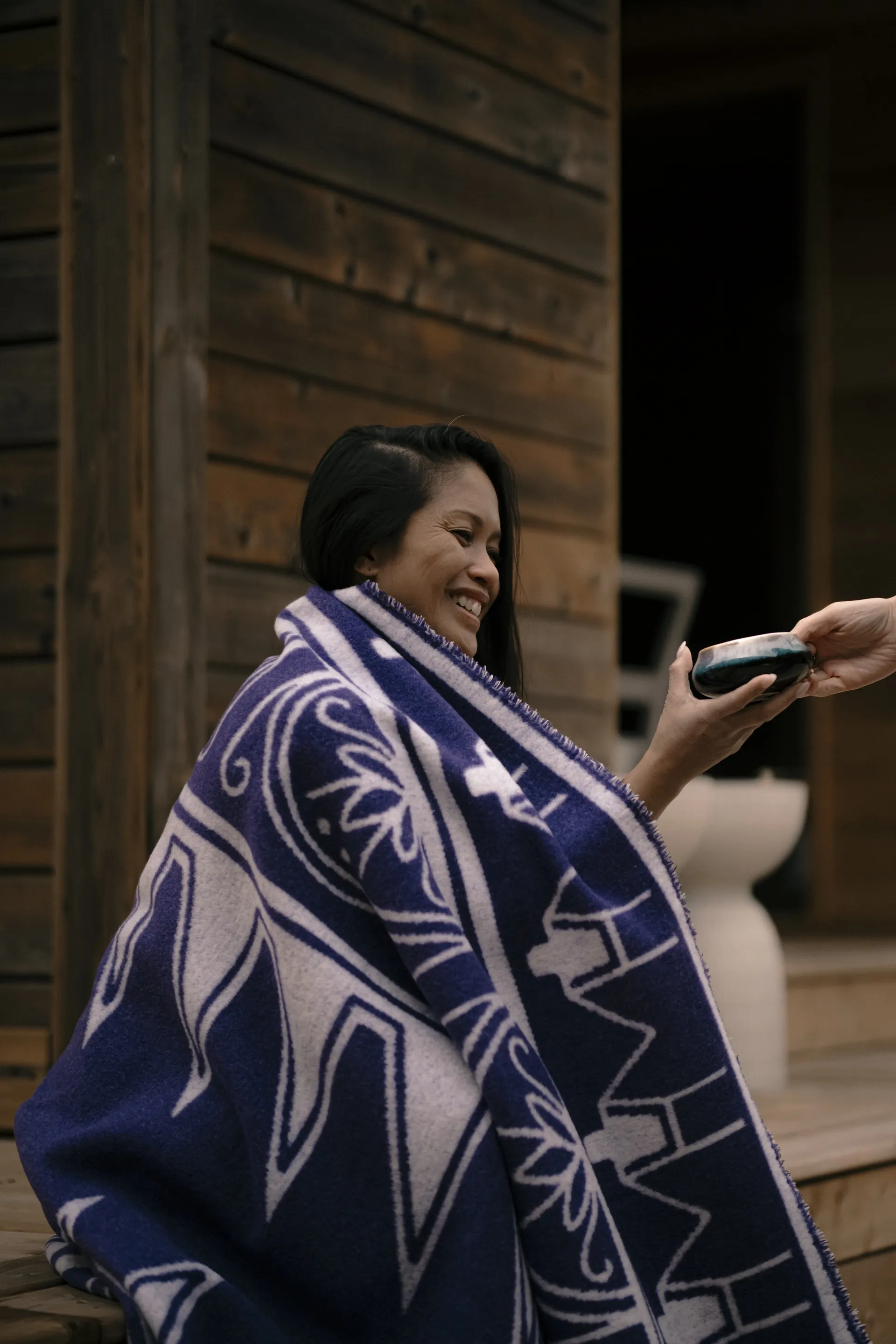 Guest wrapped in a patterned wool blanket being handed a warm bowl outside a wooden cabin