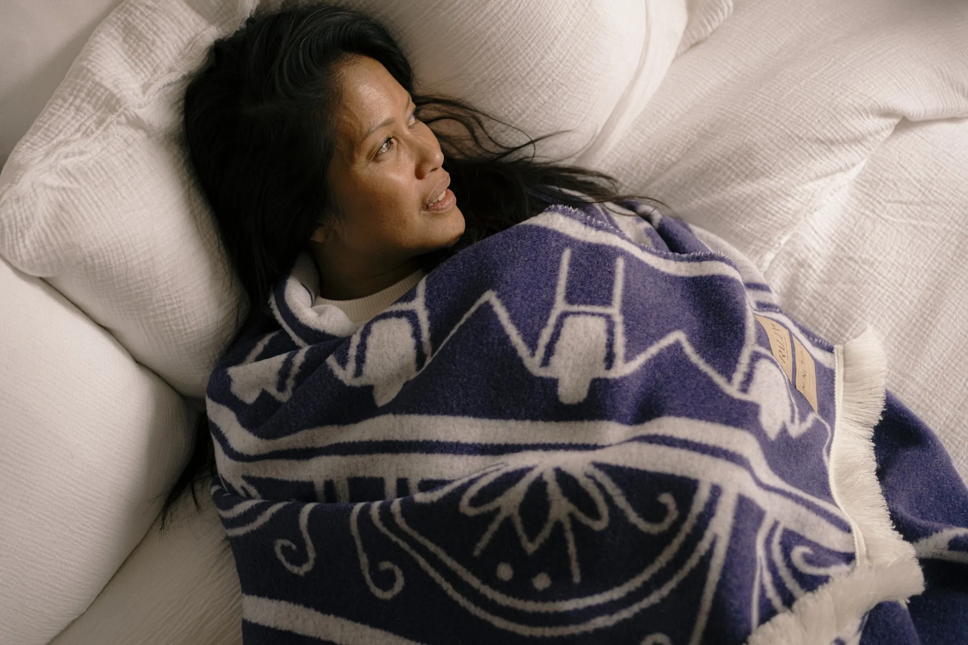 Woman resting on white bedding wrapped in a patterned navy and cream Ralia wool blanket
