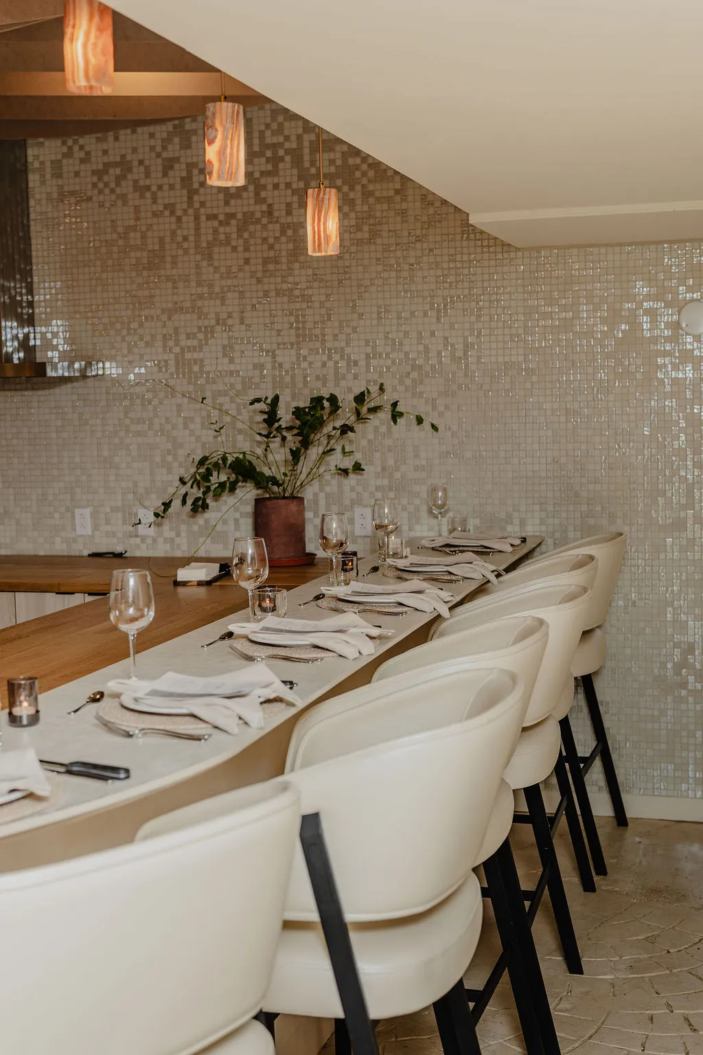 Chef's counter lined with white leather stools under wood-grain pendant lights, set for a private dinner at La Maison Ralia