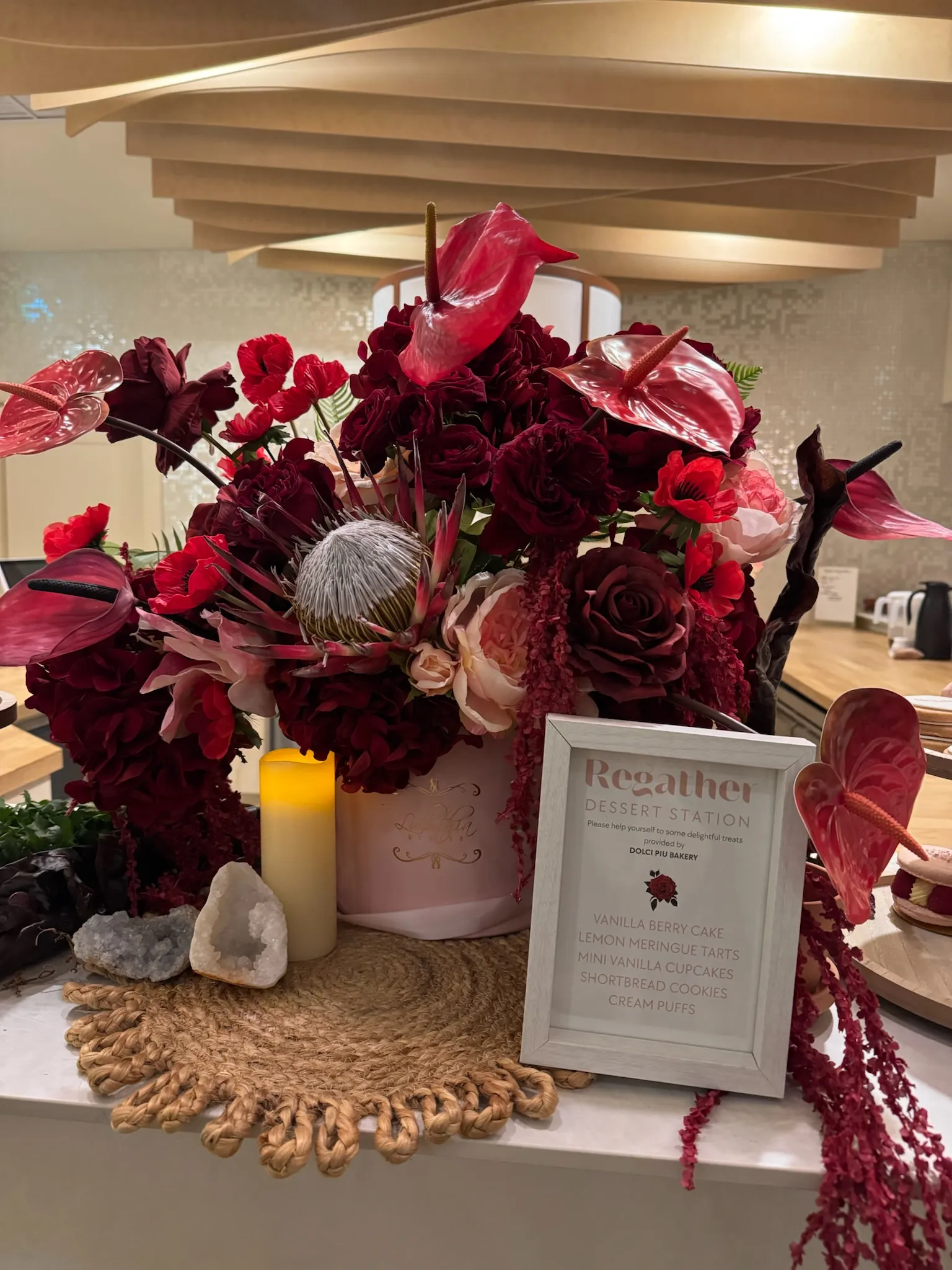 Deep red floral arrangement and framed dessert station sign beneath the wood slat ceiling of the Connections event space