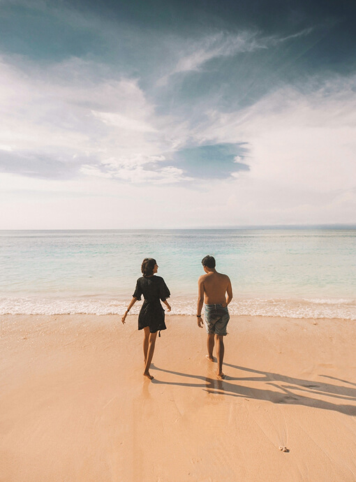 A man and a woman walk barefoot towards the turquoise ocean, on a sandy beach with gentle waves under a cloudy sky.