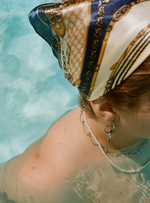 A person's head and shoulder are in bright blue water, adorned with a patterned silk headscarf, a silver hoop earring, and layered pearl and silver chain necklaces.