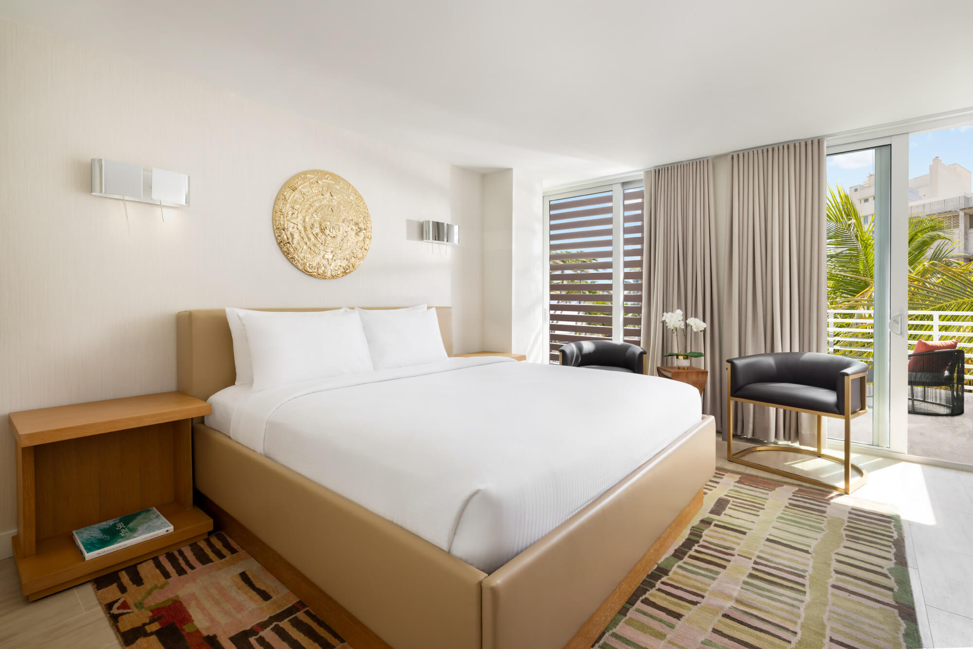 Modern hotel room with king bed, two black chairs, patterned rug, and balcony with palm view.