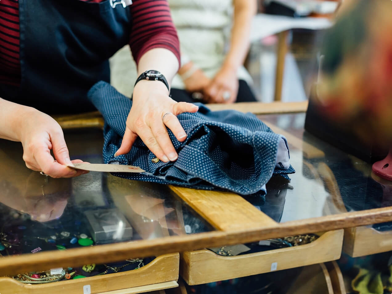 A person in a striped shirt and apron folds a dark blue polka-dotted garment, holding a tag, on a glossy counter above a jewelry display. Another person is blurred in the background.