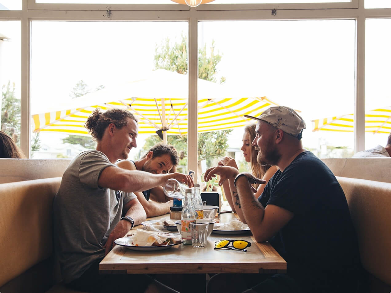 Four friends sitting in a restaurant booth with drinks and plates, chatting by a window with yellow umbrellas outside.
