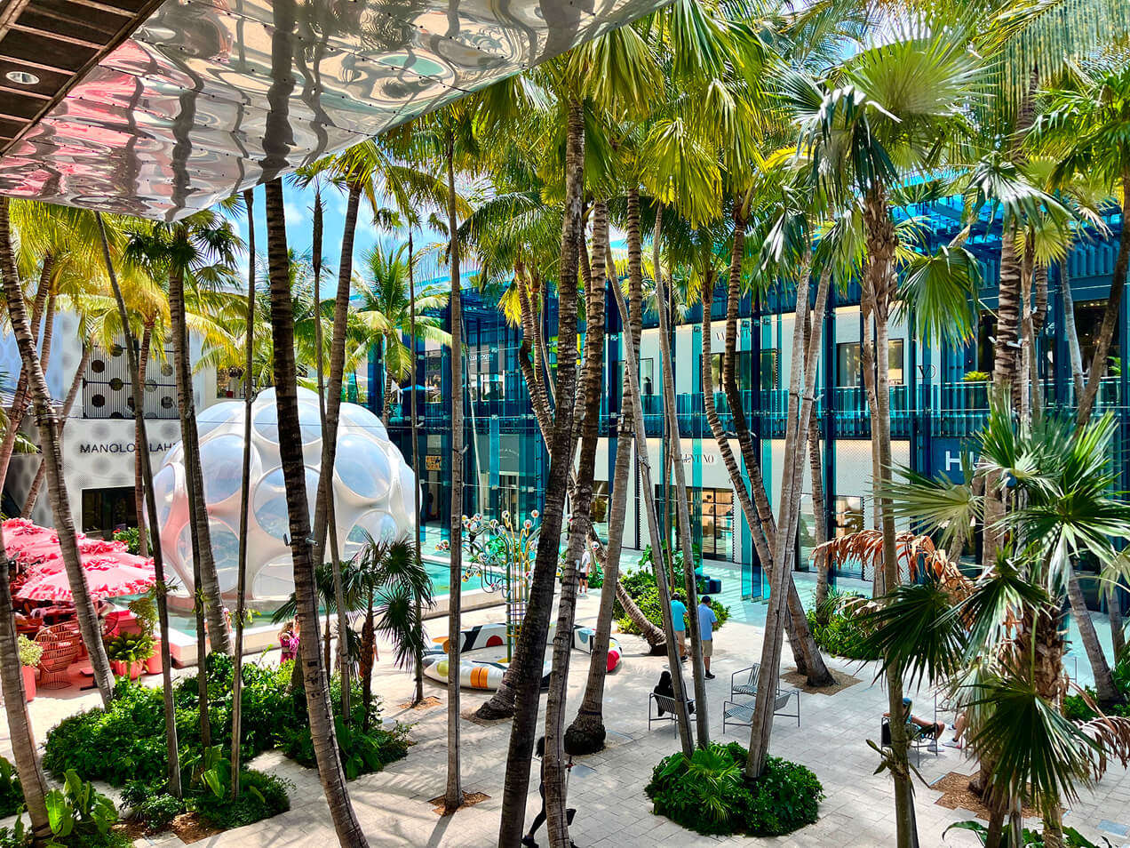 Numerous palm trees punctuate a sunlit modern shopping plaza with glass-fronted luxury stores and a spherical white structure. People relax or walk through the space. Text: MANOLO BLAHNIK, VALENTINO, LOUIS VUITTON, HUGO.