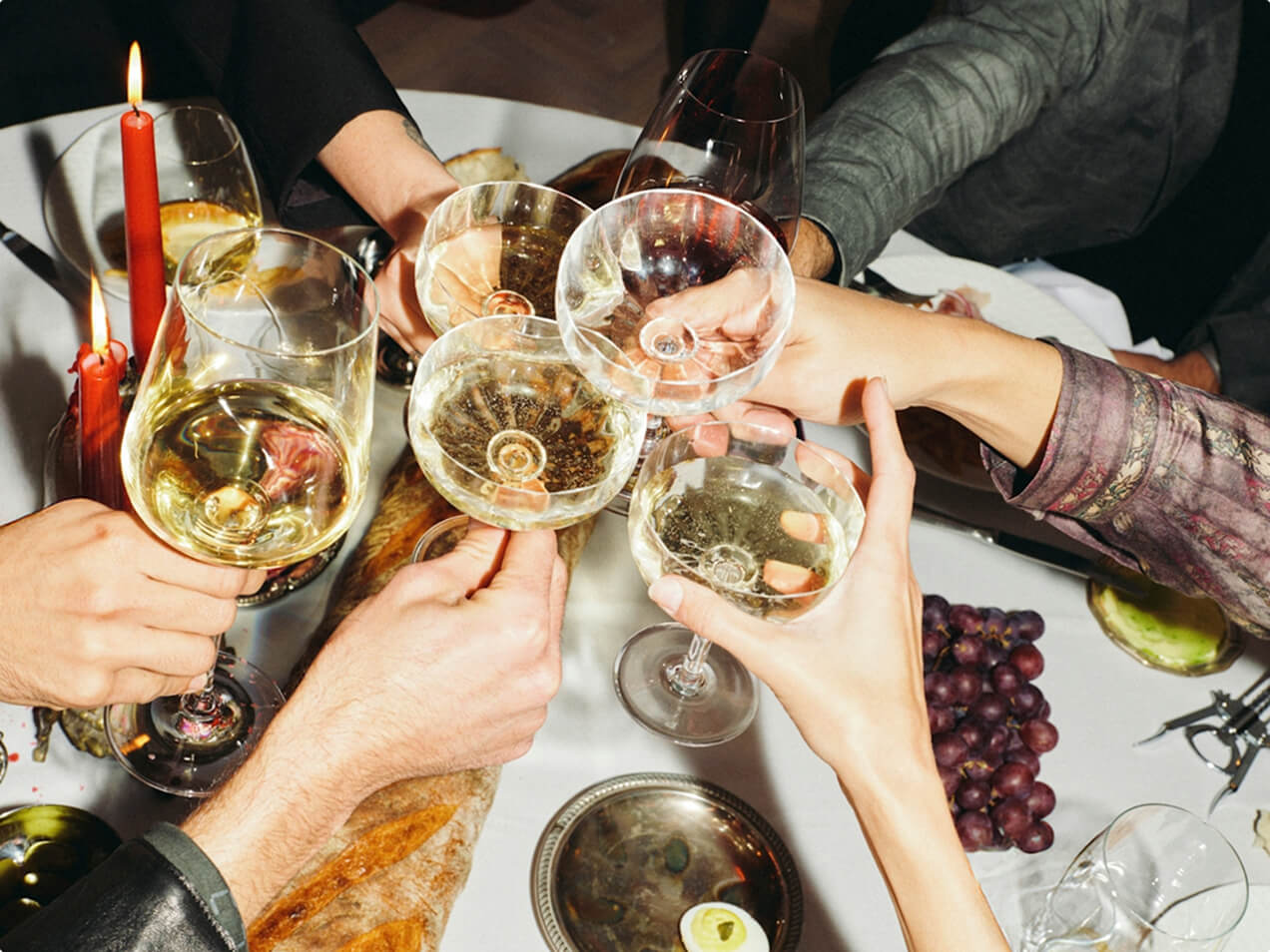 Several hands clink glasses of wine and champagne in a toast around a white tablecloth dinner table laden with baguette, grapes, and lit candles.