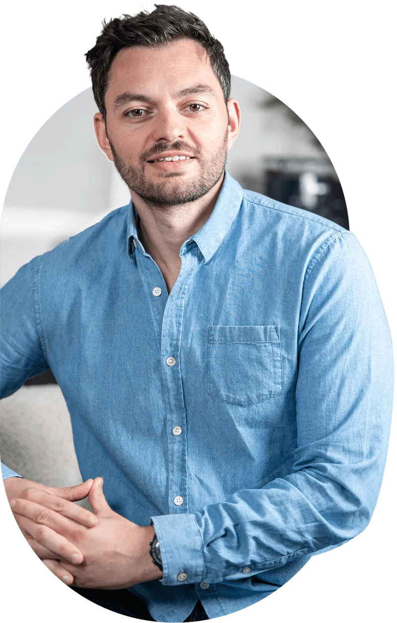 Man with short dark hair and beard wearing a light blue button-up shirt, sitting with hands clasped.