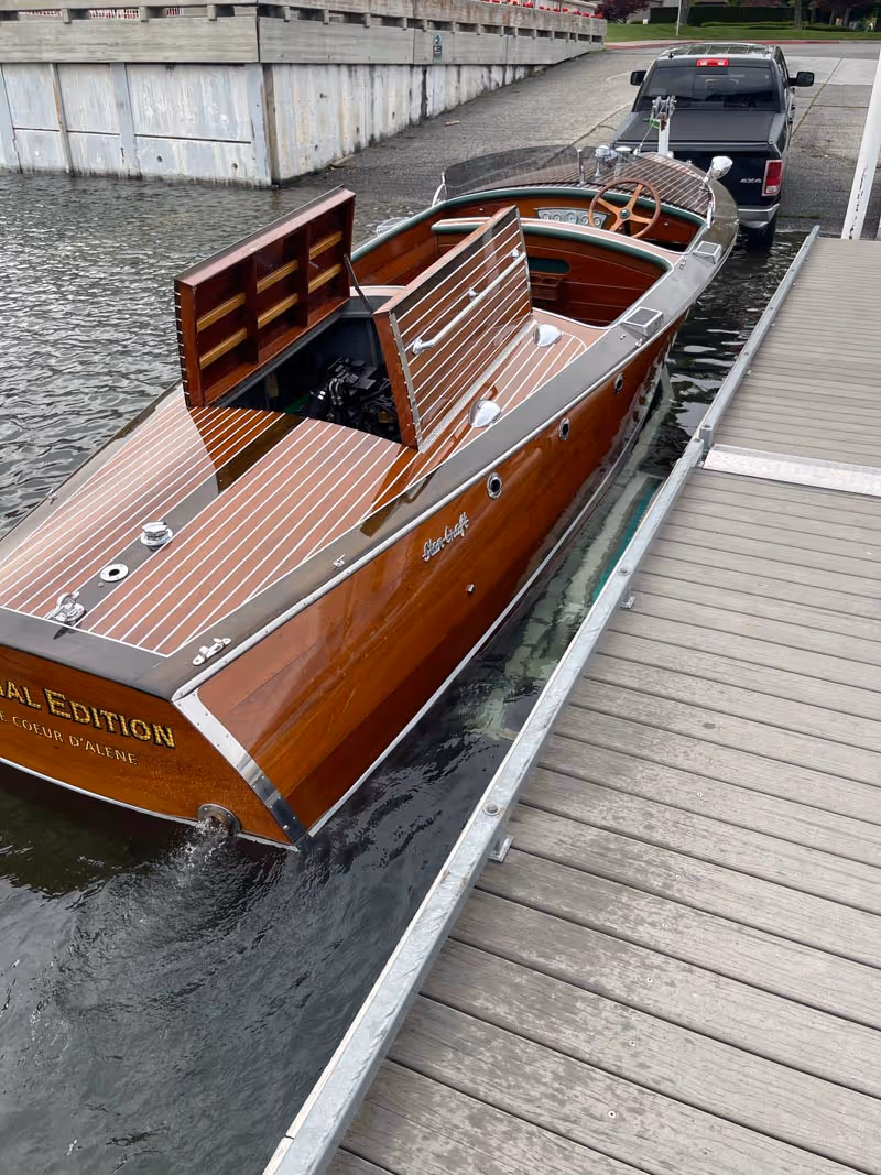 Vintage wooden motorboat with open engine hatch docked beside a wooden pier, hitched to a black pickup truck.