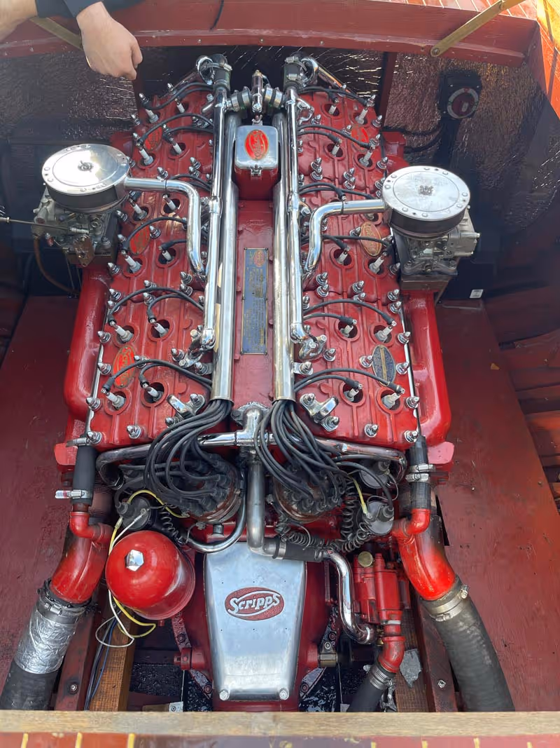 Top view of a red vintage Scripps V8 marine engine with chrome pipes and dual carburetors inside a wooden boat engine compartment.
