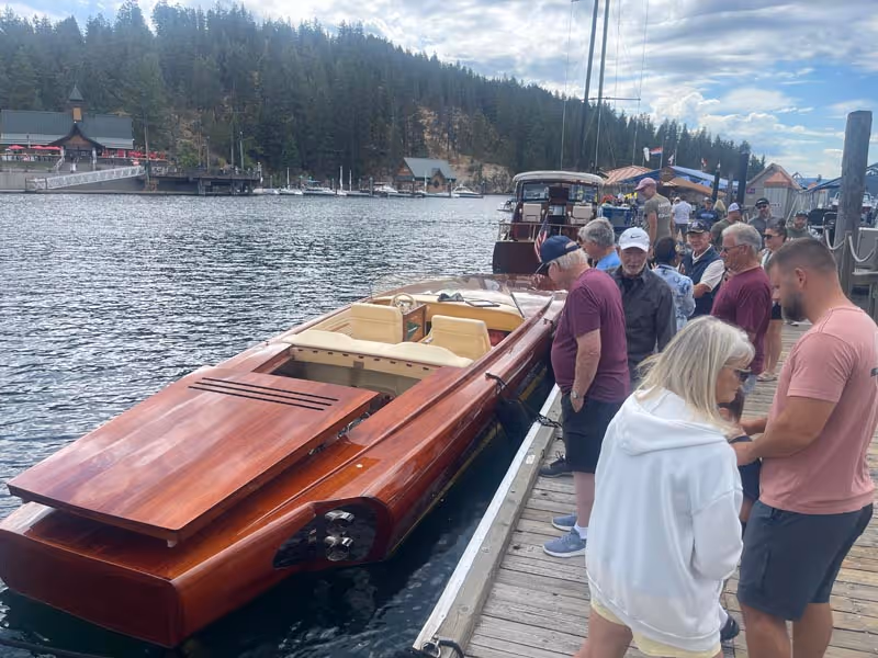 Group of people standing on a wooden dock around a sleek wooden speedboat on a lake with forested hills in the background.