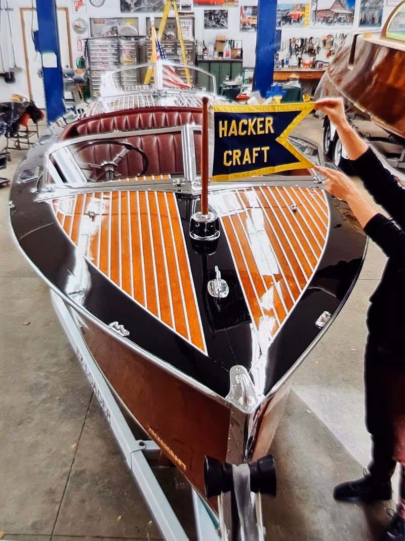 Shiny wooden motorboat with polished deck and maroon leather seating in a workshop, featuring a flag that reads 'Hacker Craft'.