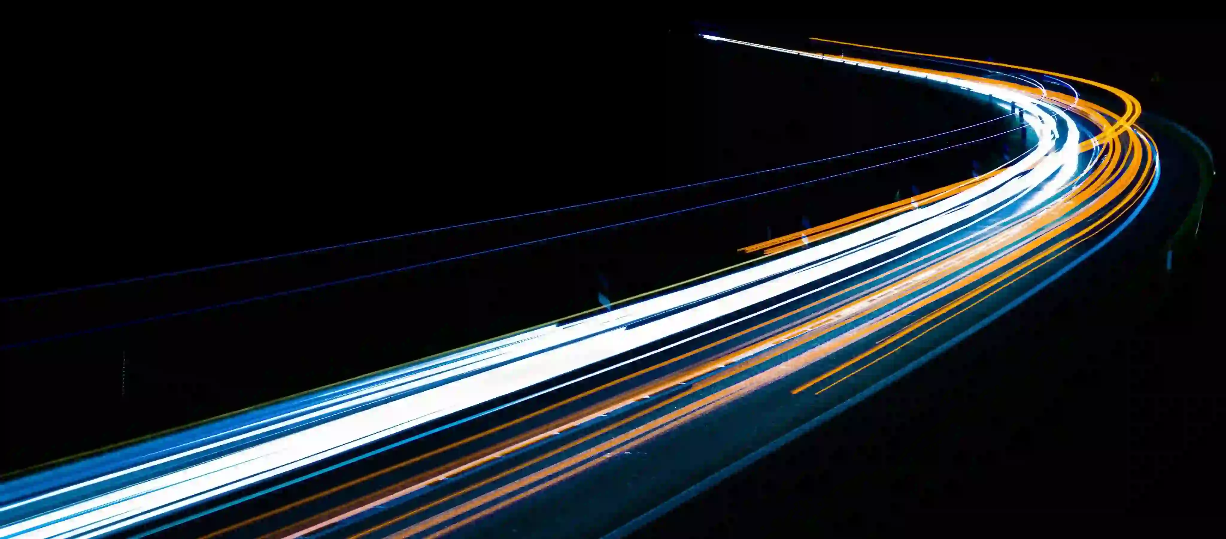 Light trails of vehicles on a curved road at night showing white and orange streaks.