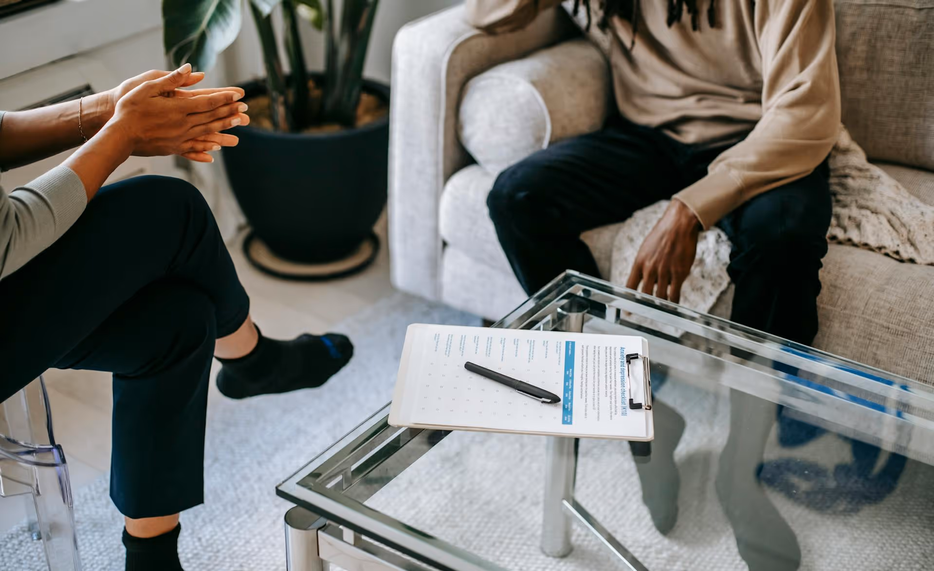 Two people having a conversation in a living room with a clipboard and pen on a glass coffee table between them.