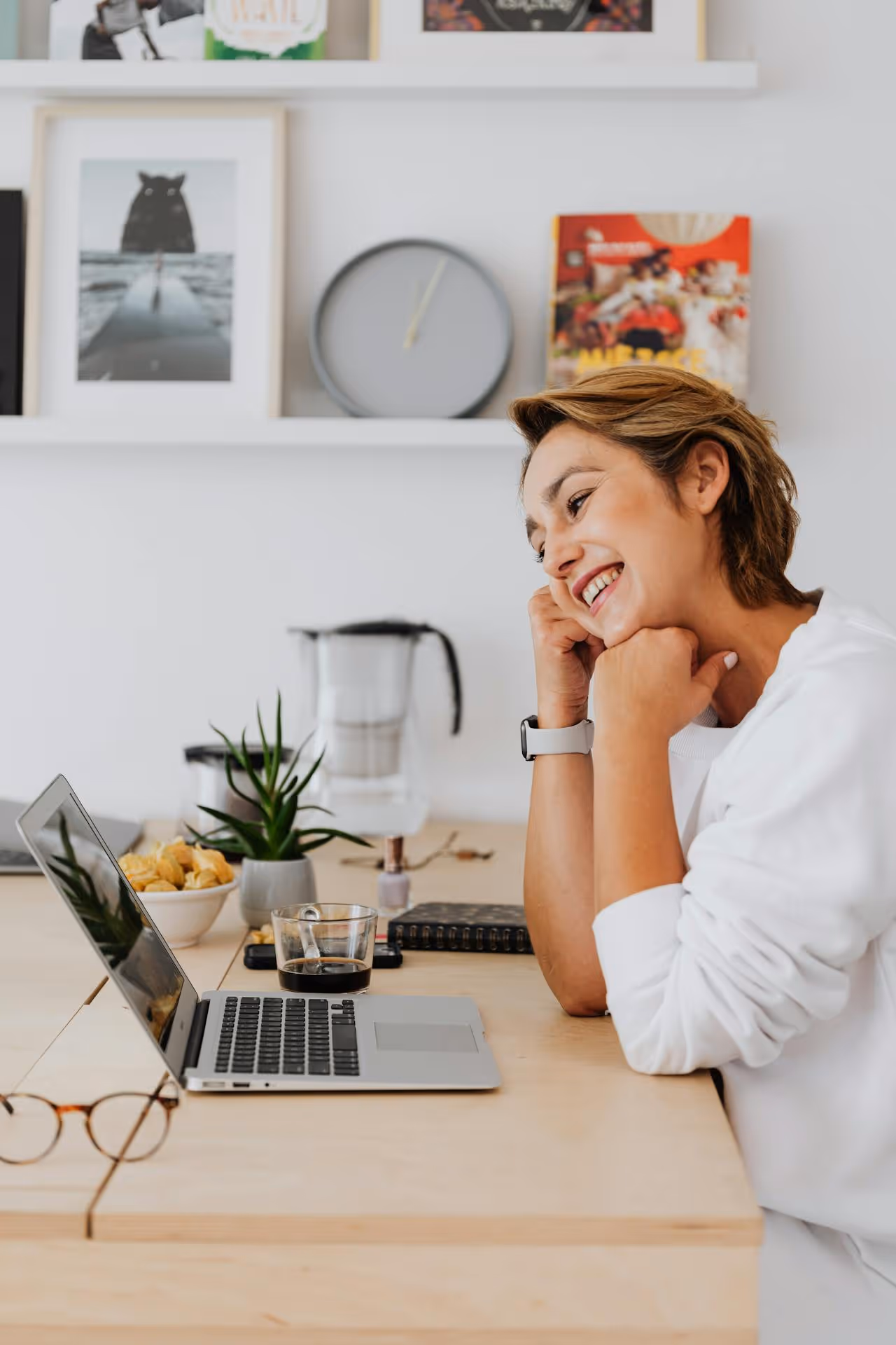 Smiling woman in a white sweater sitting at a wooden desk looking at a laptop screen with coffee and snacks nearby.