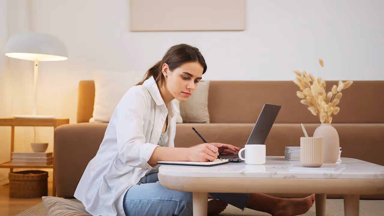 Woman sitting on the floor writing in a notebook with a laptop on a coffee table in a cozy living room.