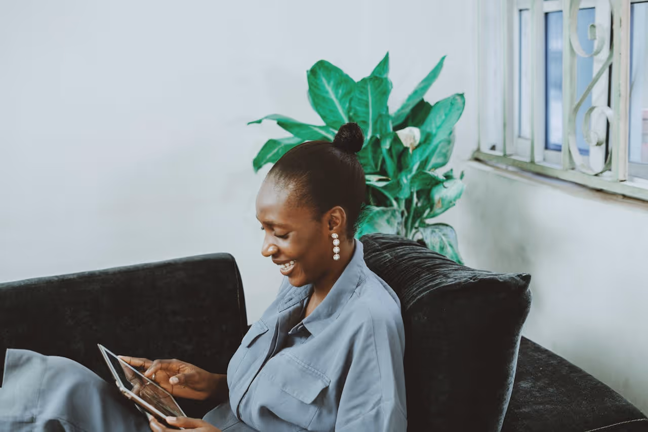 Smiling woman sitting on a black couch using a tablet with a green plant and window in the background.