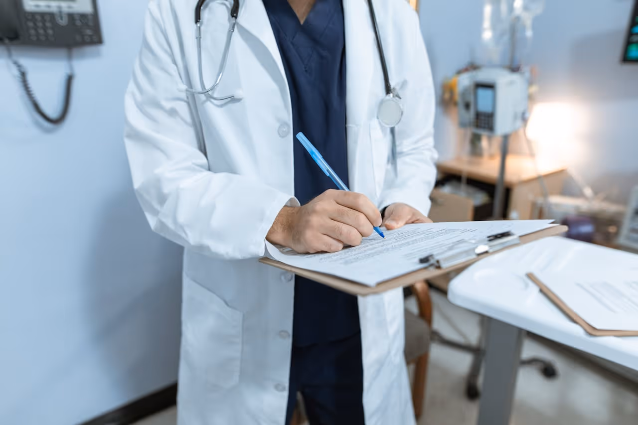 Doctor in a white coat writing notes on a clipboard in a medical office.