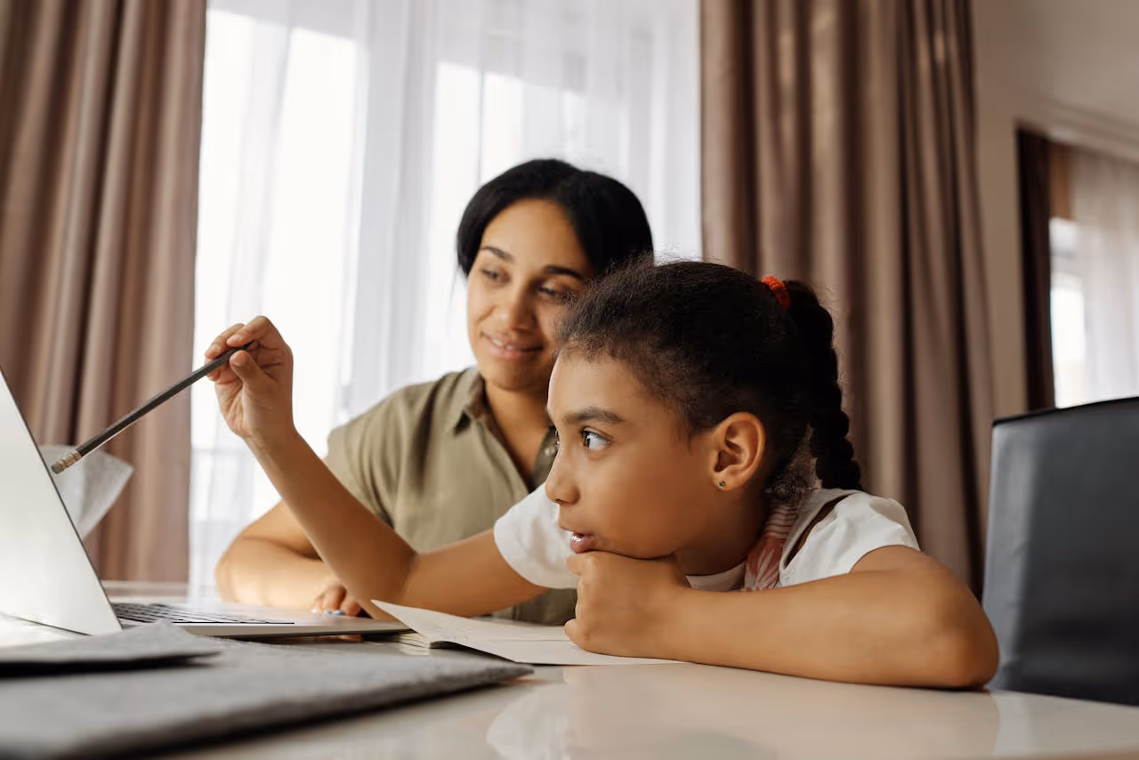 Young girl pointing with a pencil at a laptop screen while an adult woman watches and smiles in a home setting.