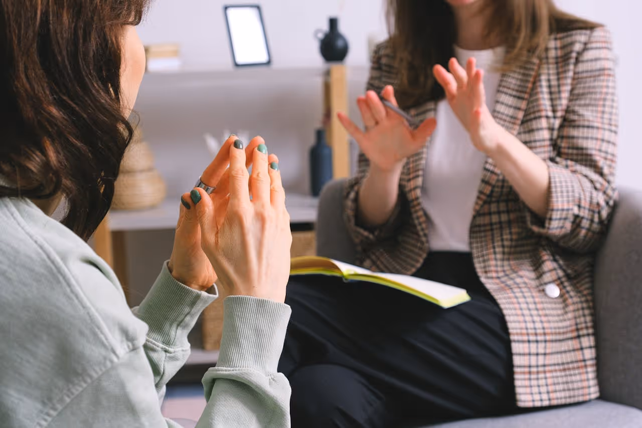 Two women engaged in a conversation, one wearing a plaid blazer holds a pen and notebook on her lap.