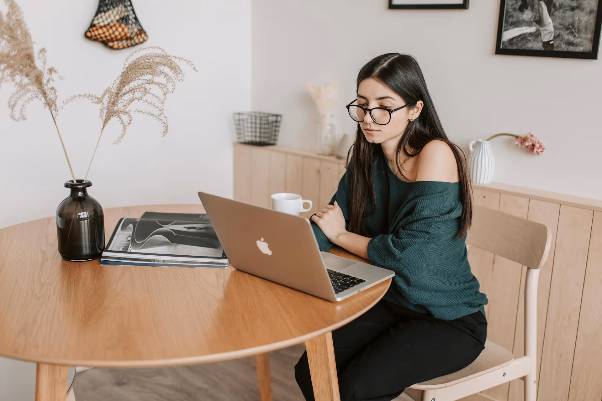 Young woman with glasses sitting at a wooden table working on a laptop in a cozy minimalist room.
