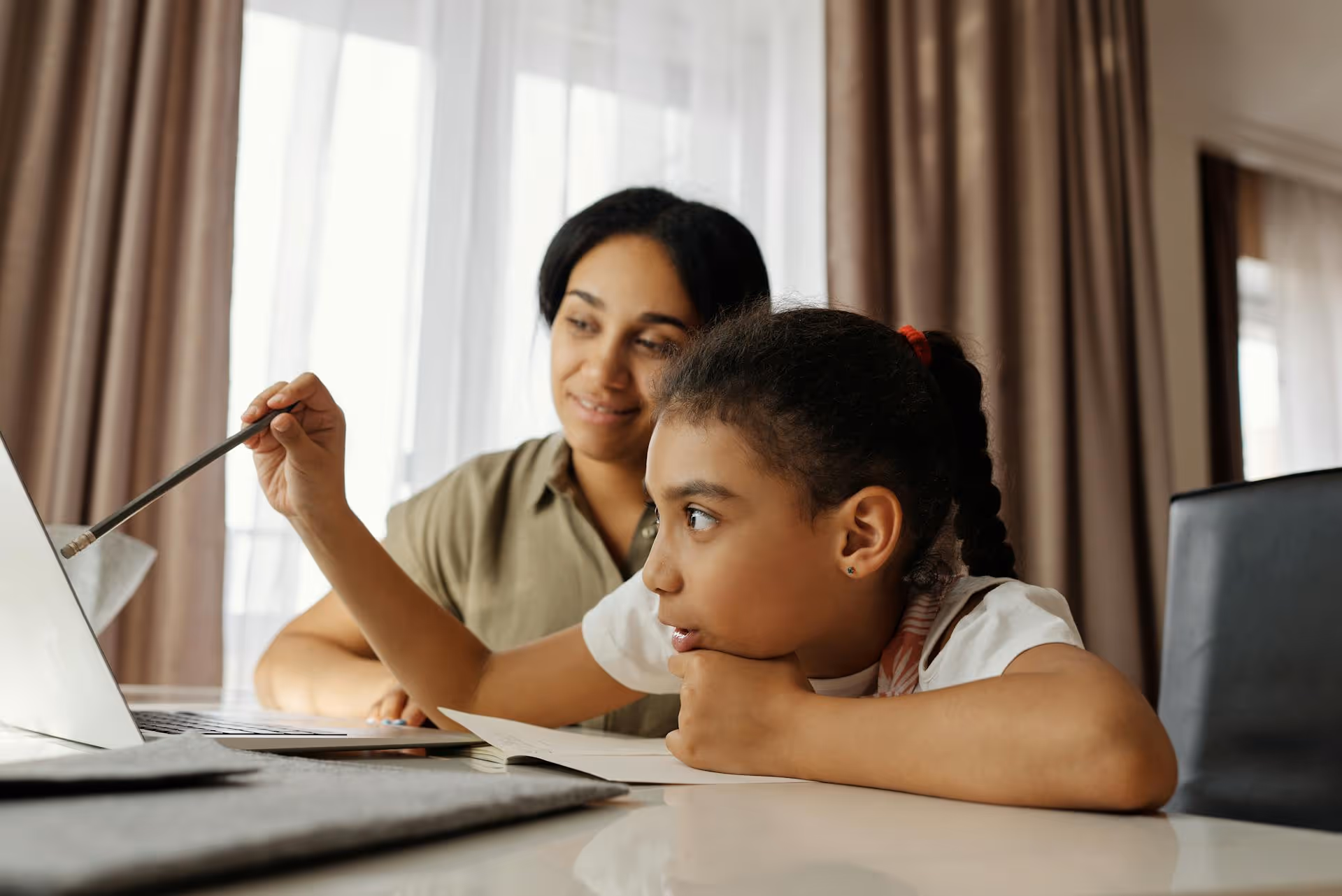 Young girl with braided hair pointing at a laptop screen with a pencil while an adult woman watches and smiles.