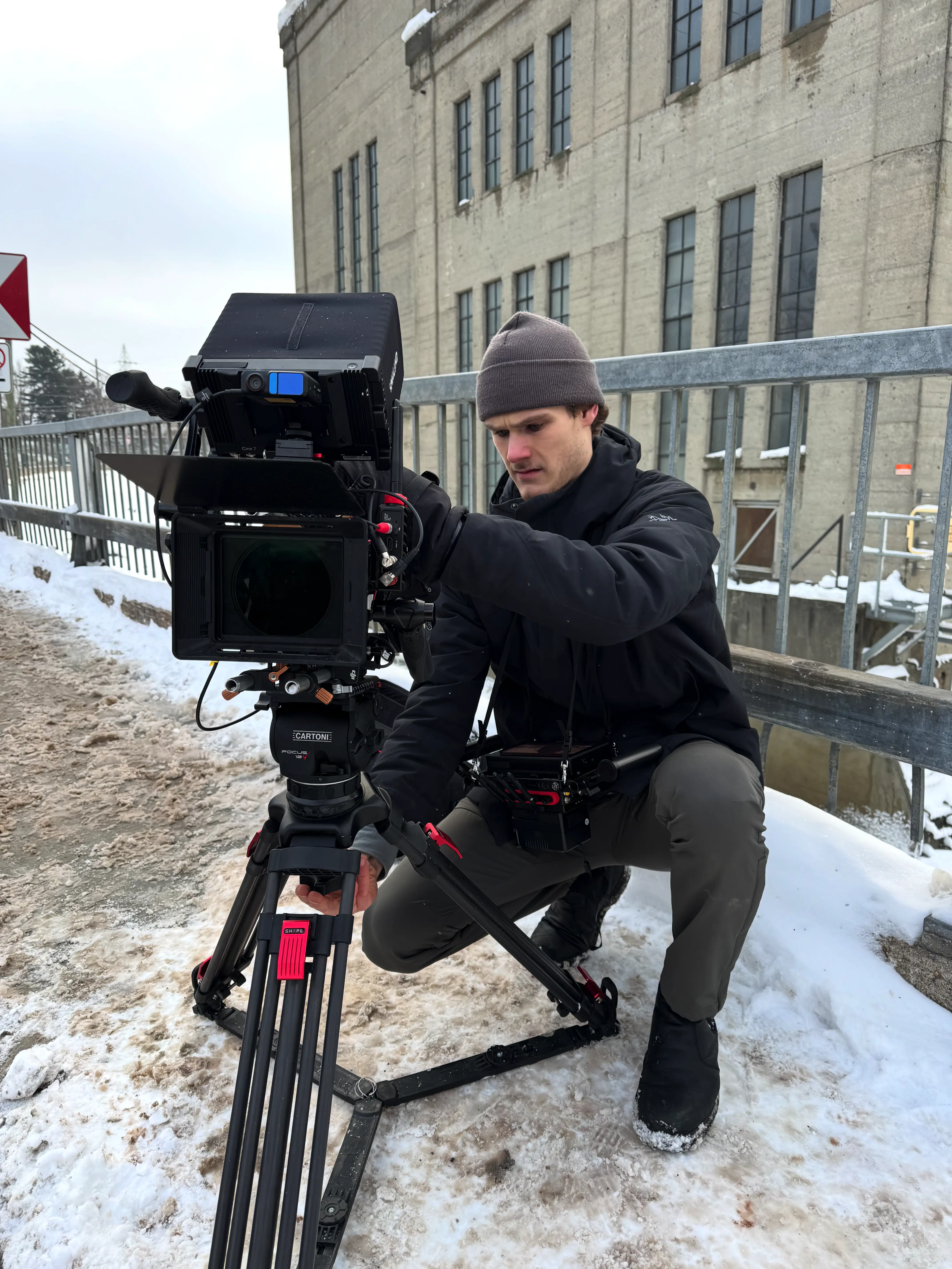 Justin Desjardins accroupi ajuste une caméra professionnelle sur un trépied dans la neige devant un bâtiment.