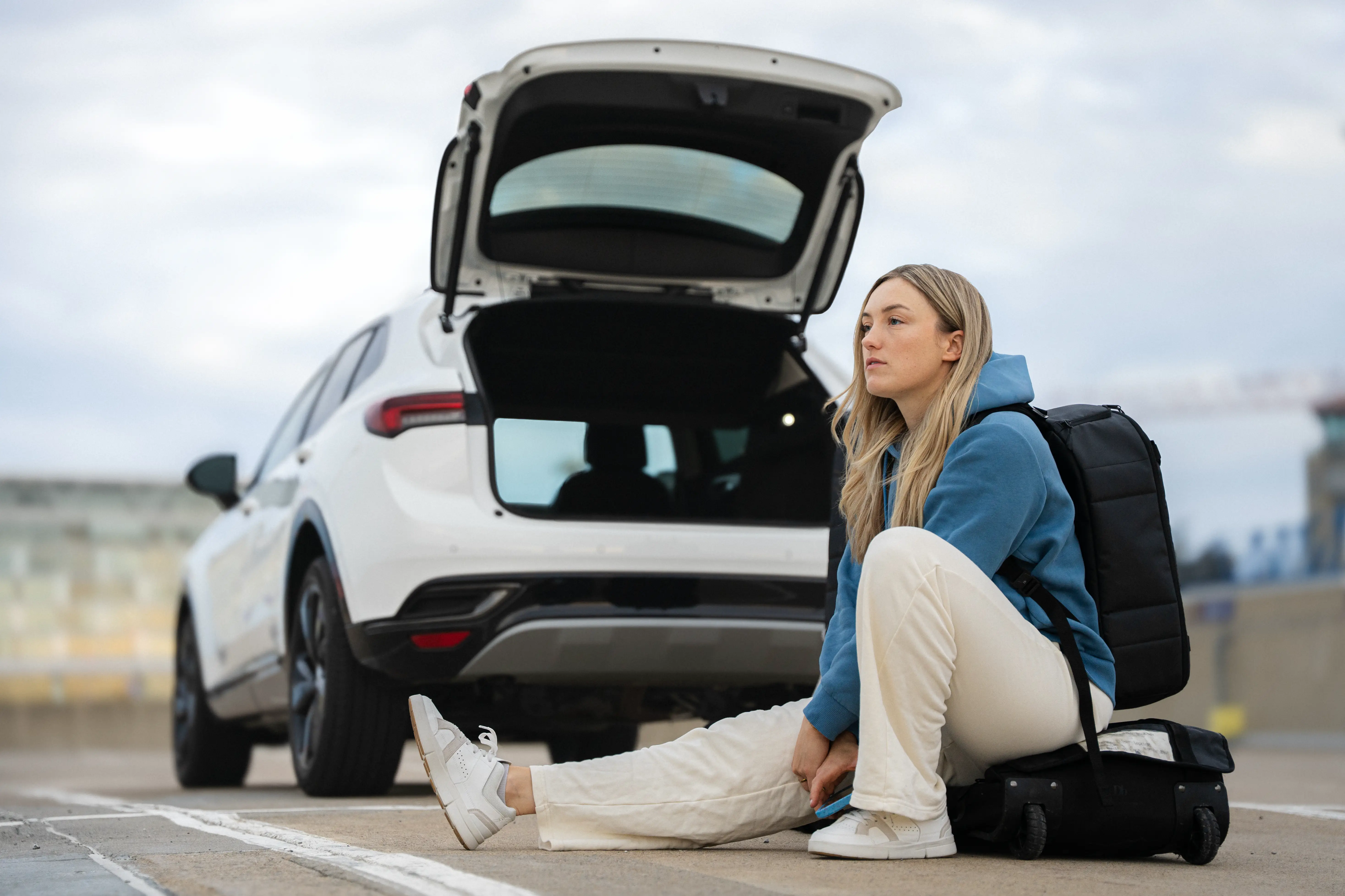 Jeune femme blonde en sweat bleu assise sur une valise devant une voiture blanche avec le coffre ouvert.