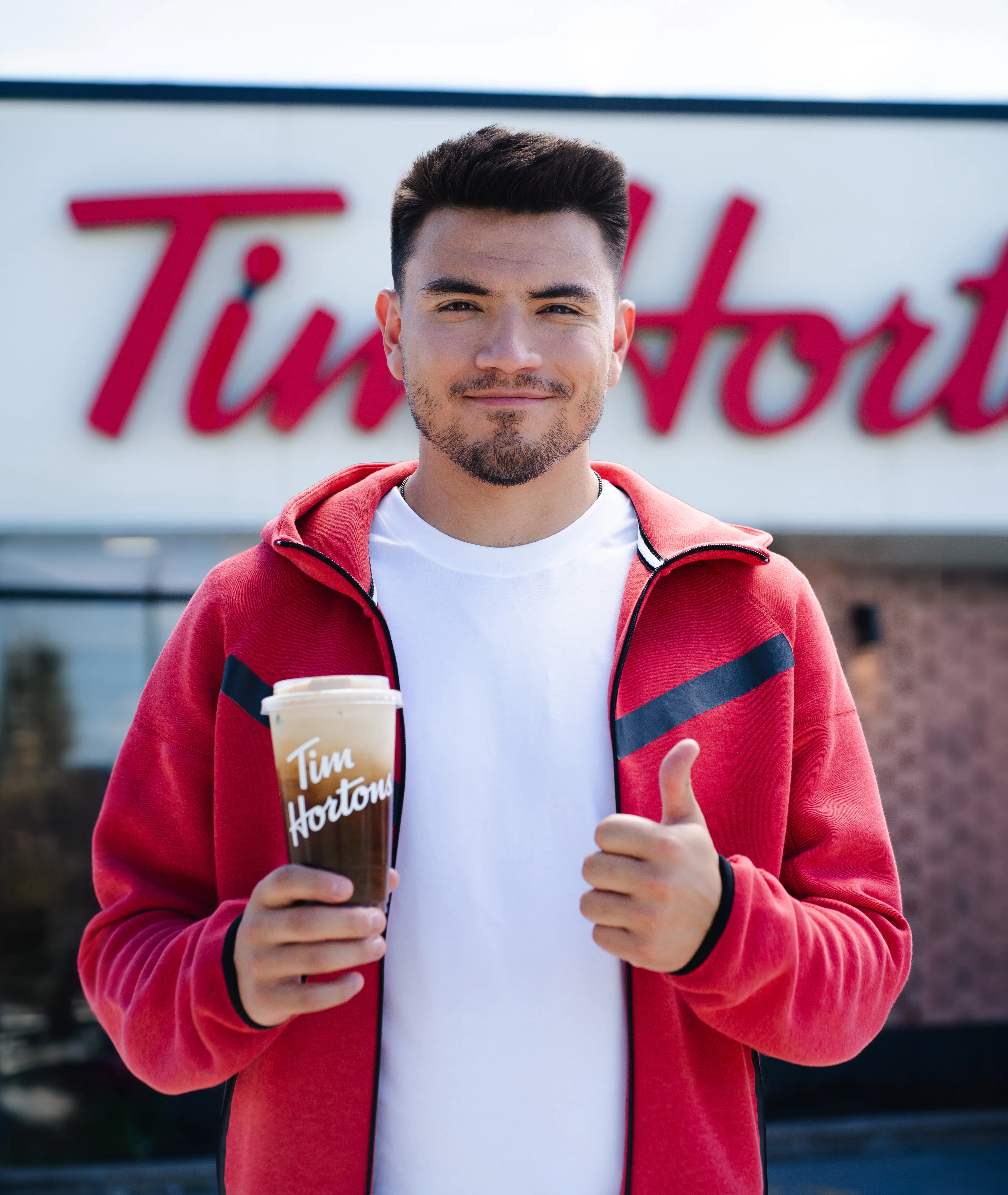 Nick Suzuki souriant en veste rouge tenant une boisson Tim Hortons et faisant un pouce levé.