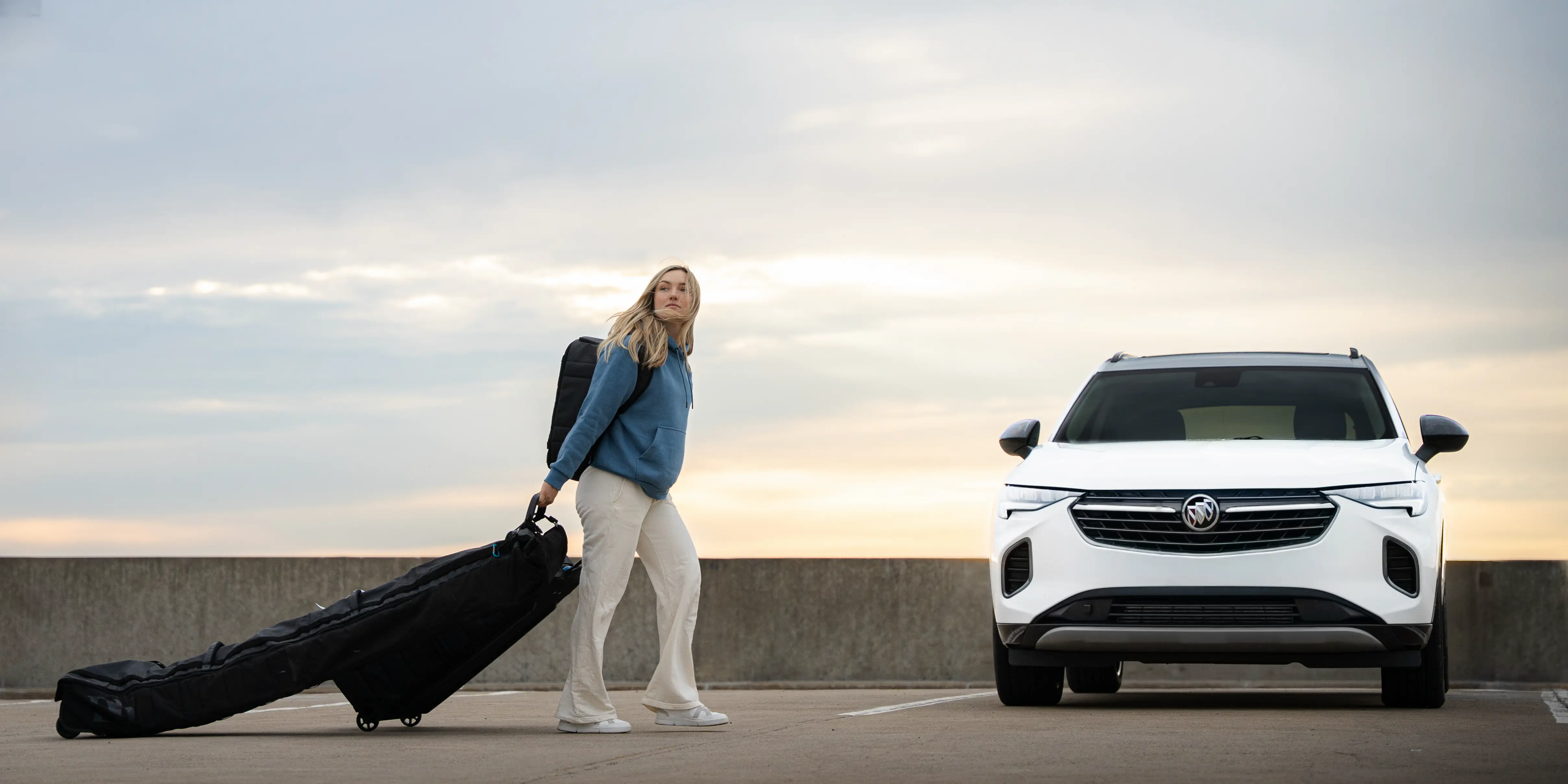 Femme avec sac à dos tirant une valise roulettes vers un VUS blanc garé au coucher du soleil.
