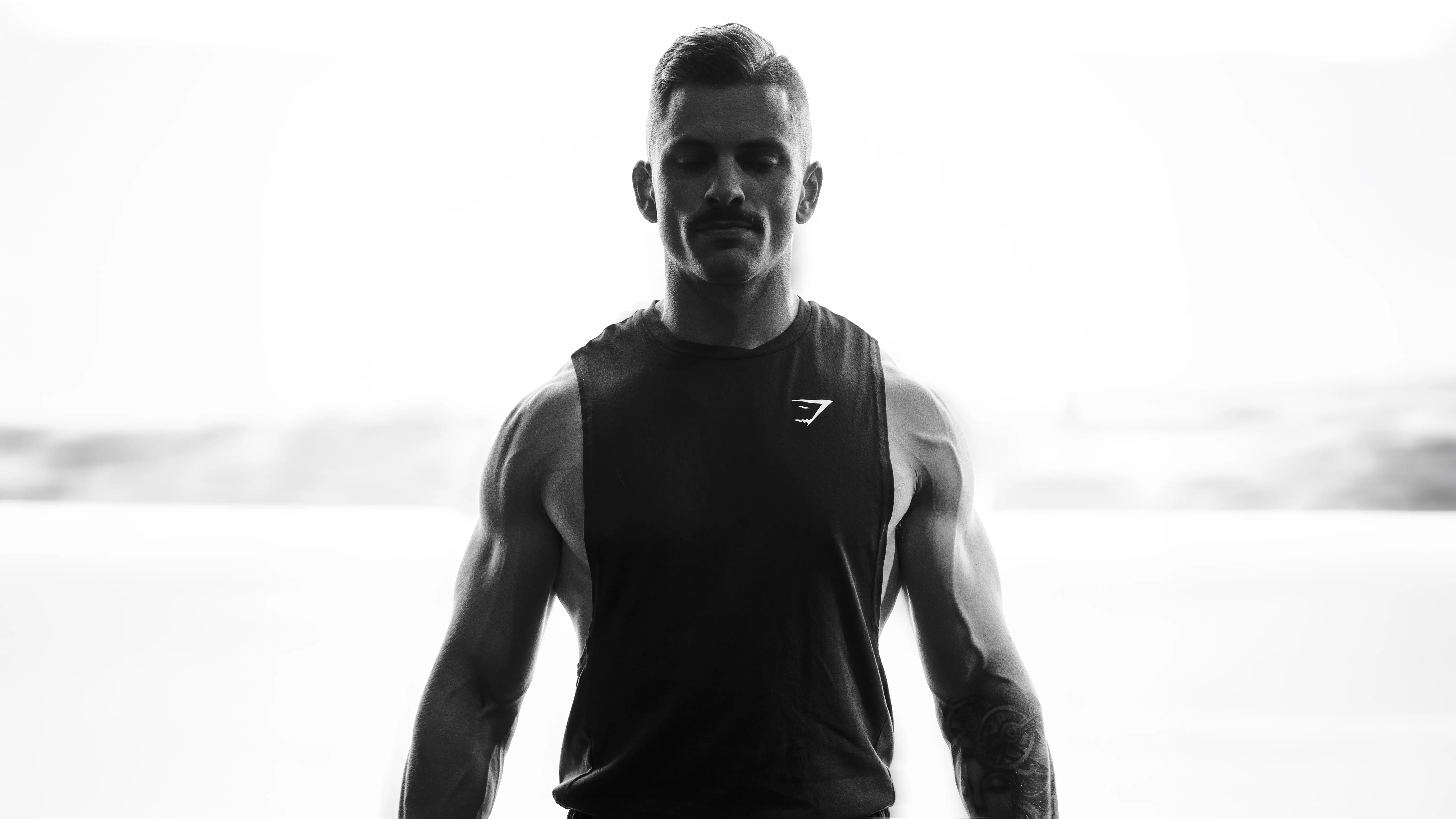 Homme musclé en débardeur noir Gymshark, yeux fermés, fond lumineux flou.
