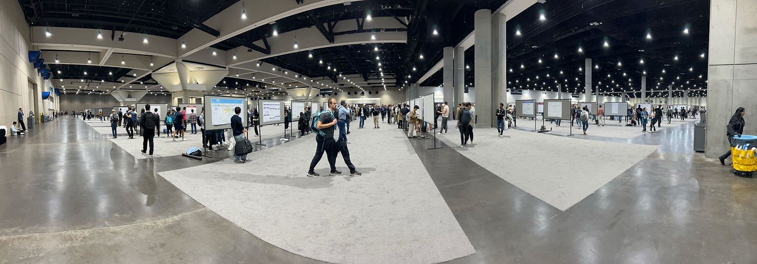 Spacious convention hall with people viewing posters on display boards. Some walk around, others read. Bright lighting, gray flooring.