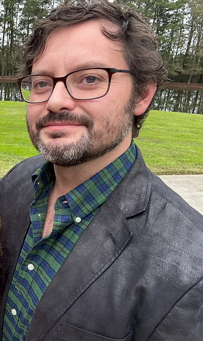 Randy Davila, with glasses and beard in a leather jacket and green plaid shirt, standing outdoors near a grassy area and trees, smiling.
