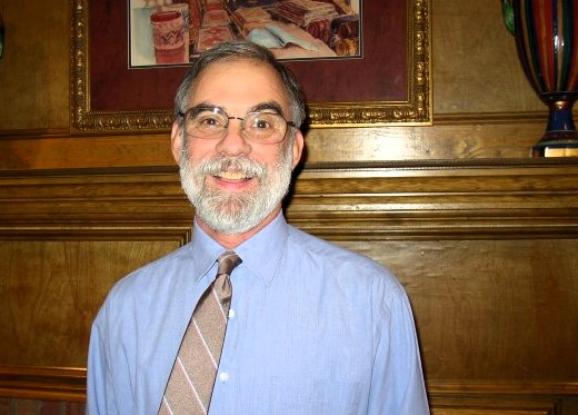 Bill Staton, in blue collared shirt with tie standing in front of wooden wall indoors.