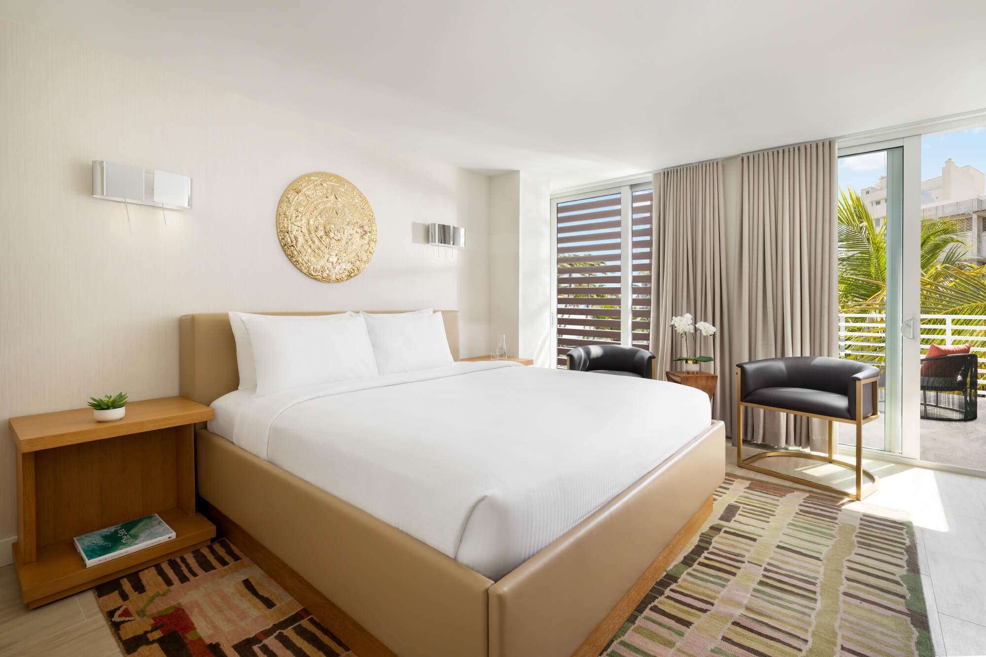 beds with white linen and tan frames are positioned side-by-side in a bright room with golden wall art. A sliding door opens to a sunny balcony.