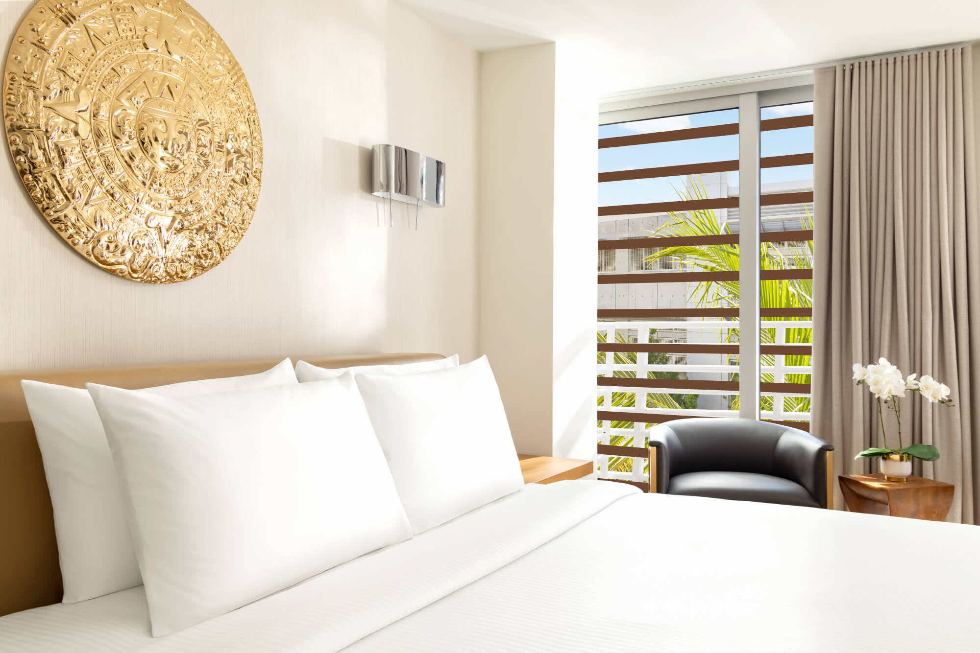 Two beds with white linen and tan frames are positioned side-by-side in a bright room with golden wall art. A sliding door opens to a balcony with palm trees.