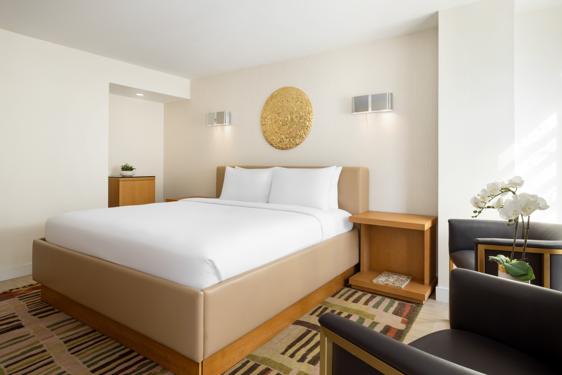 Two neatly made beds with white linens furnish a modern hotel room. Golden circular art hangs above the beds on light walls, beside a wooden cabinet and dresser.