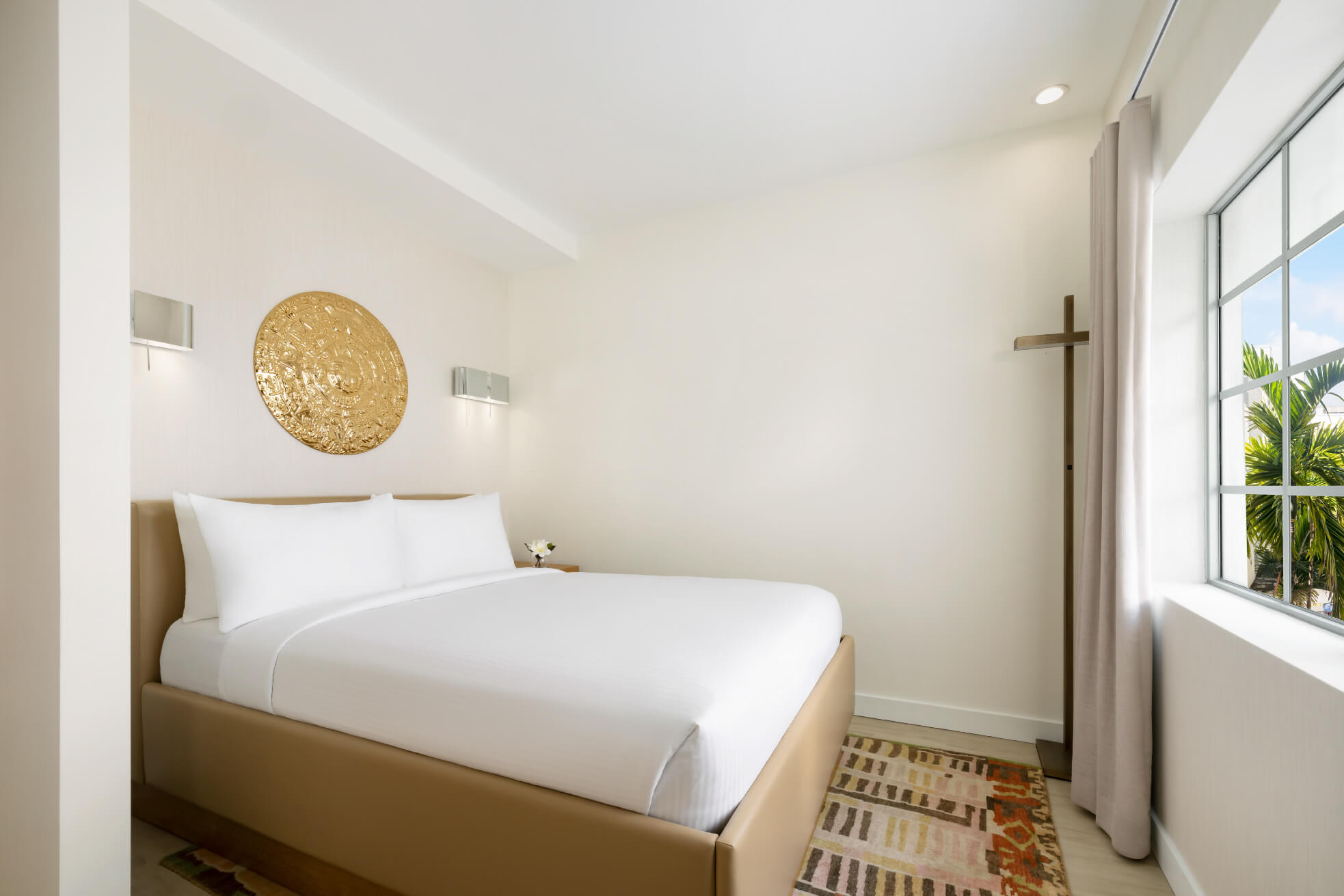 Bed with white linen and tan frames are positioned side-by-side in a bright room with golden wall art. Window view with palm trees.