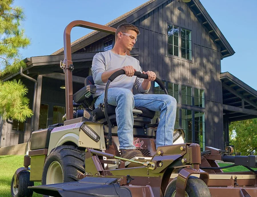 Operator on a Grasshopper zero-turn mower outside a dark wood-sided house