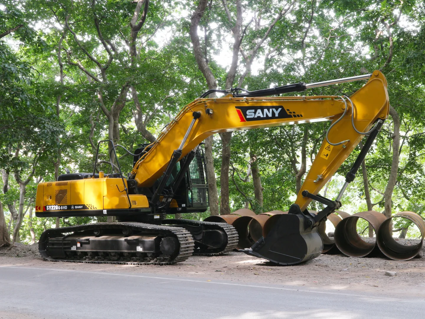 SANY SY220C crawler excavator parked under trees beside steel pipe sections