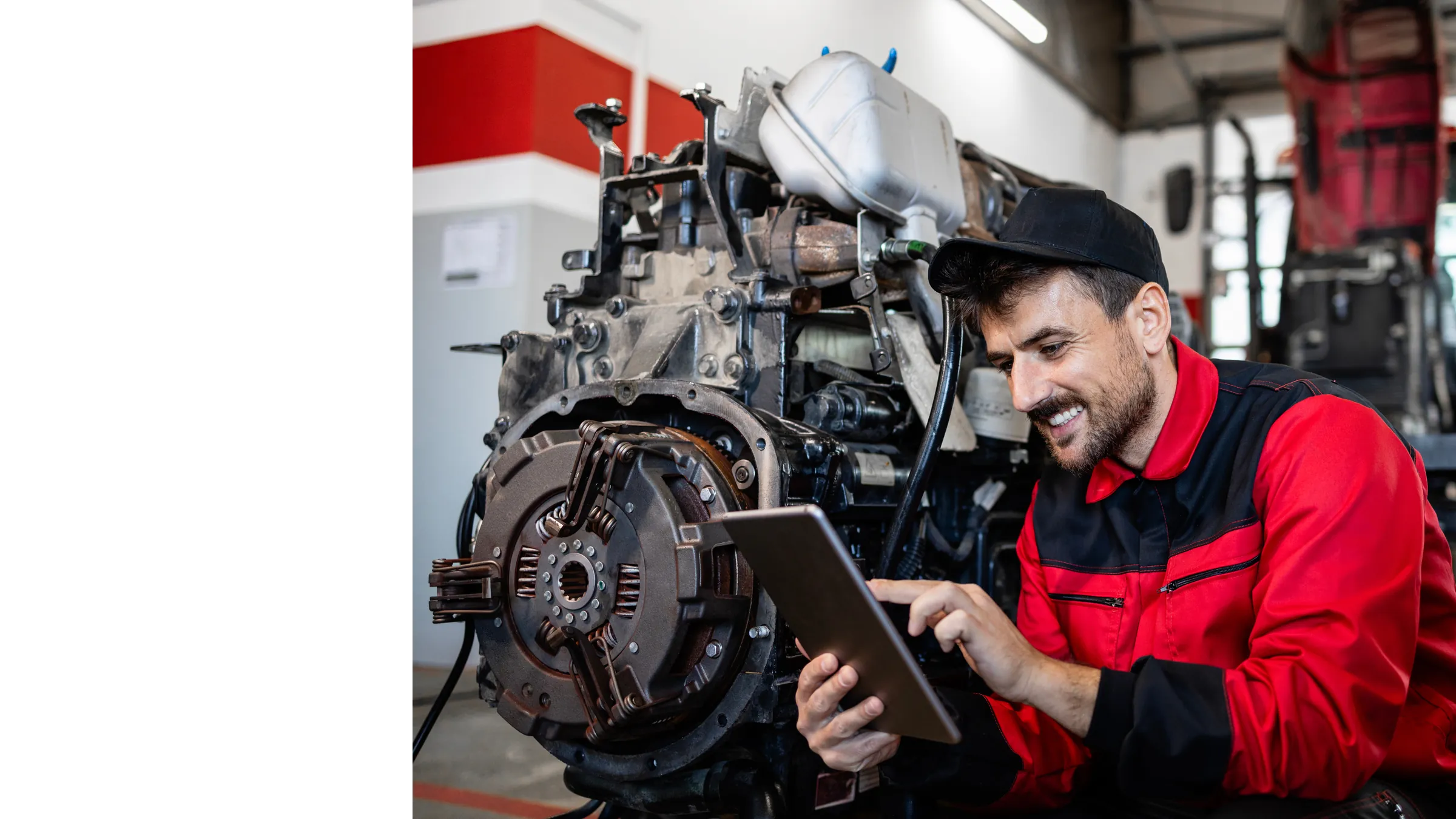 Smiling technician in red coveralls checking a work order on a tablet beside an engine in the service shop