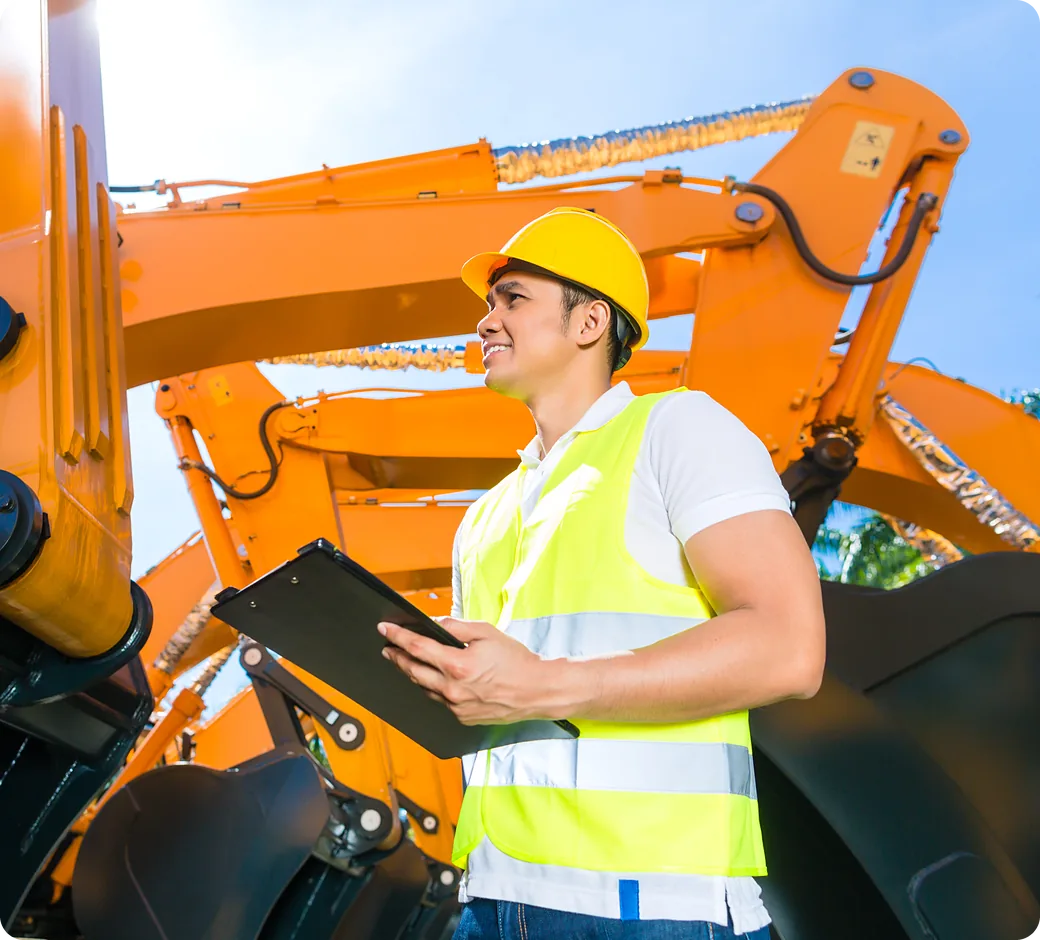 Yard worker in a hard hat and safety vest checks a tablet beside an excavator