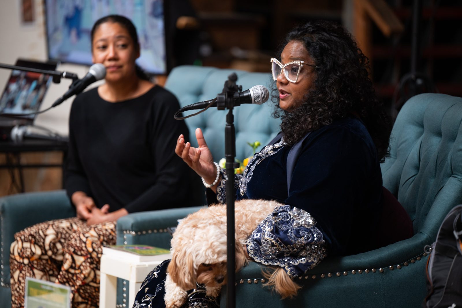 Two people seated in armchairs on stage at East Bay Meditation Center, speaking into microphones during a conversation, one of them mid-gesture with a small dog on her lap