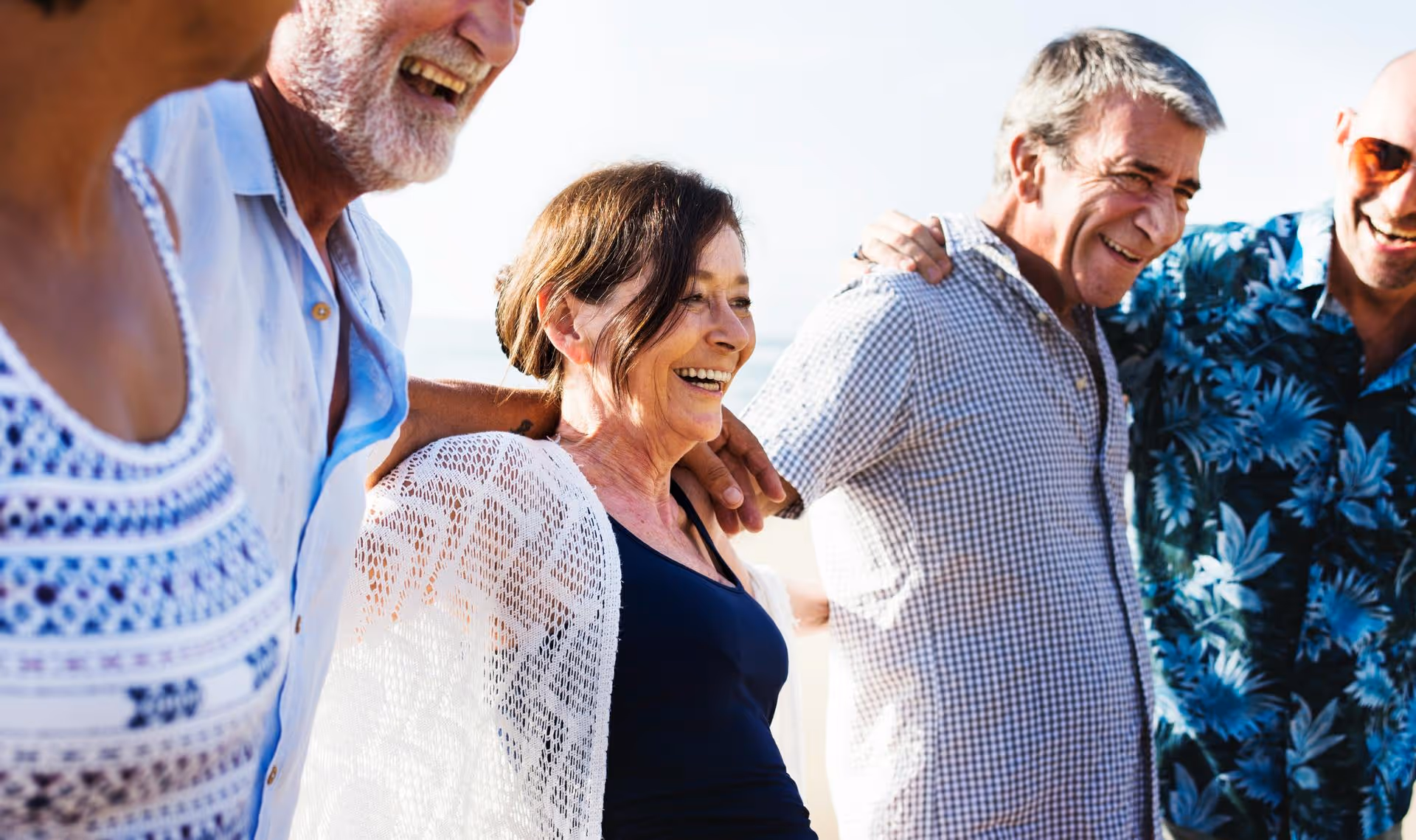 Group of four middle-aged adults standing close with arms around each other, smiling and laughing outdoors in sunlight.