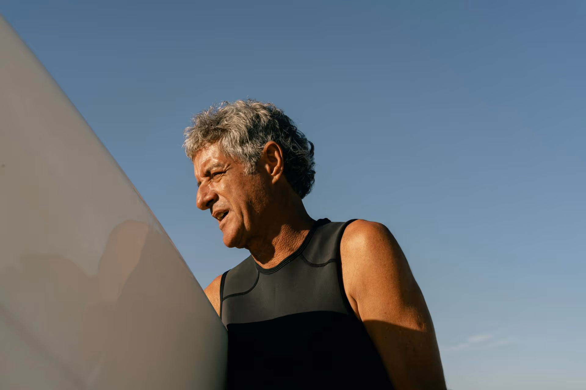 Older man with gray hair wearing a sleeveless black wetsuit holding a white surfboard against a clear blue sky.