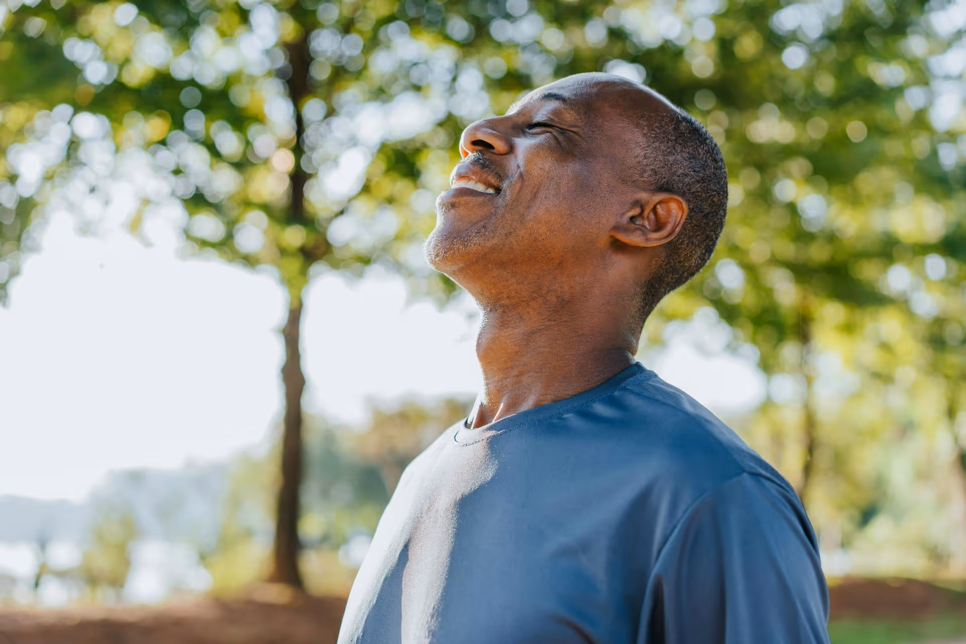 Smiling middle-aged man with eyes closed, enjoying a sunny day outdoors with trees in the background.