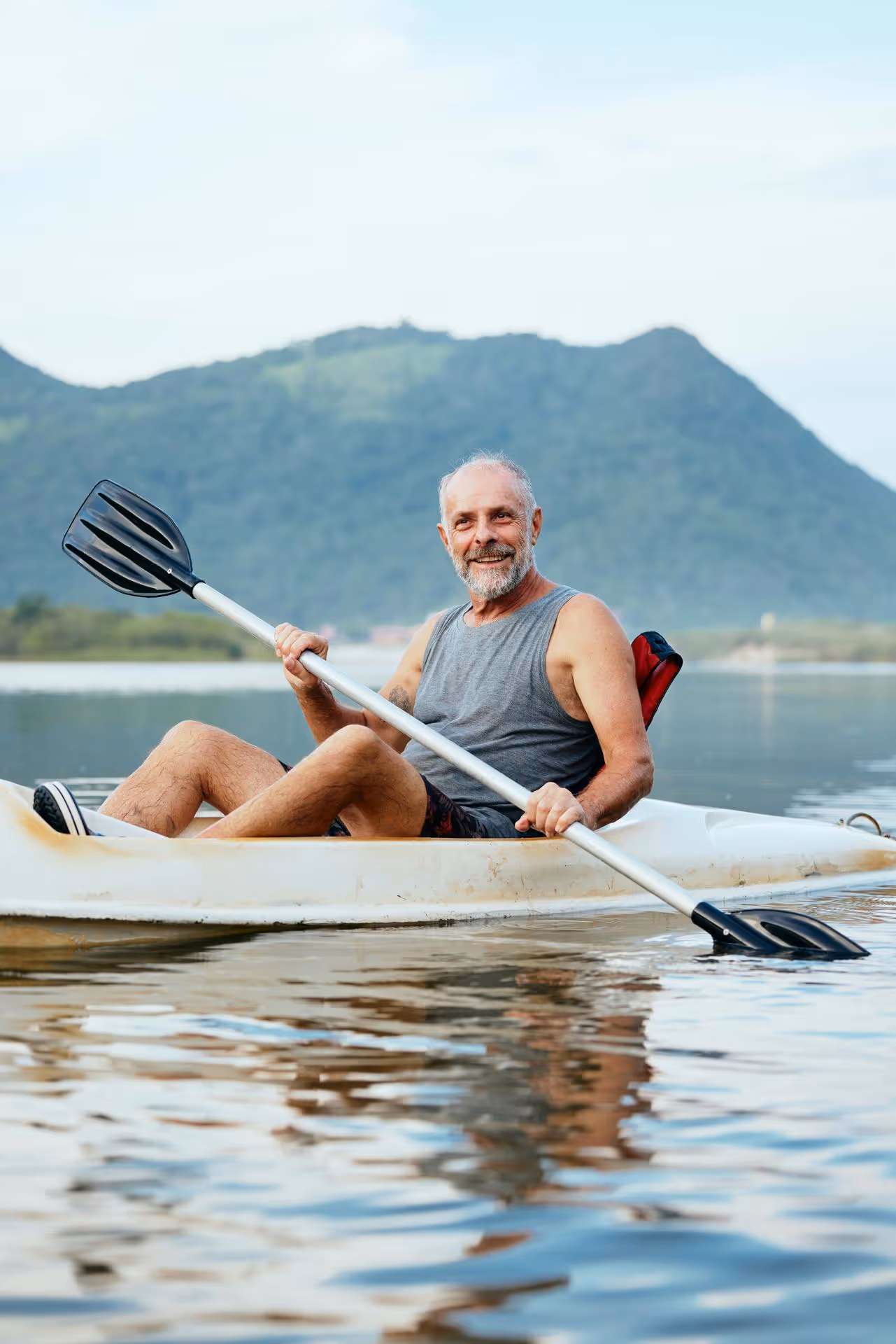 Smiling older man paddling a kayak on calm water with a green mountain background.