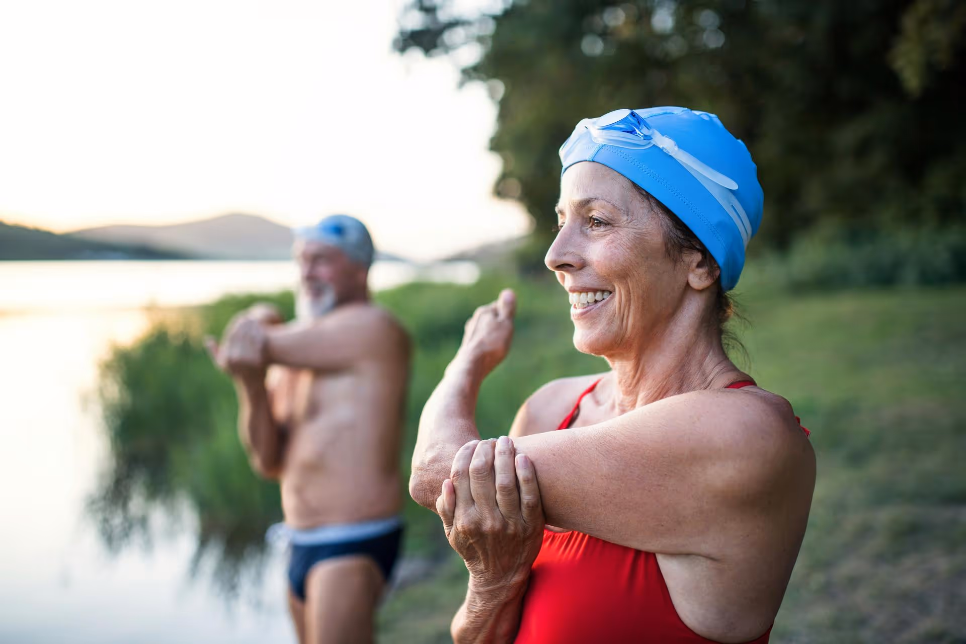 Smiling older woman in a red swimsuit and blue swim cap stretching her arm outdoors near a lake at sunset, with a blurred man stretching in the background.