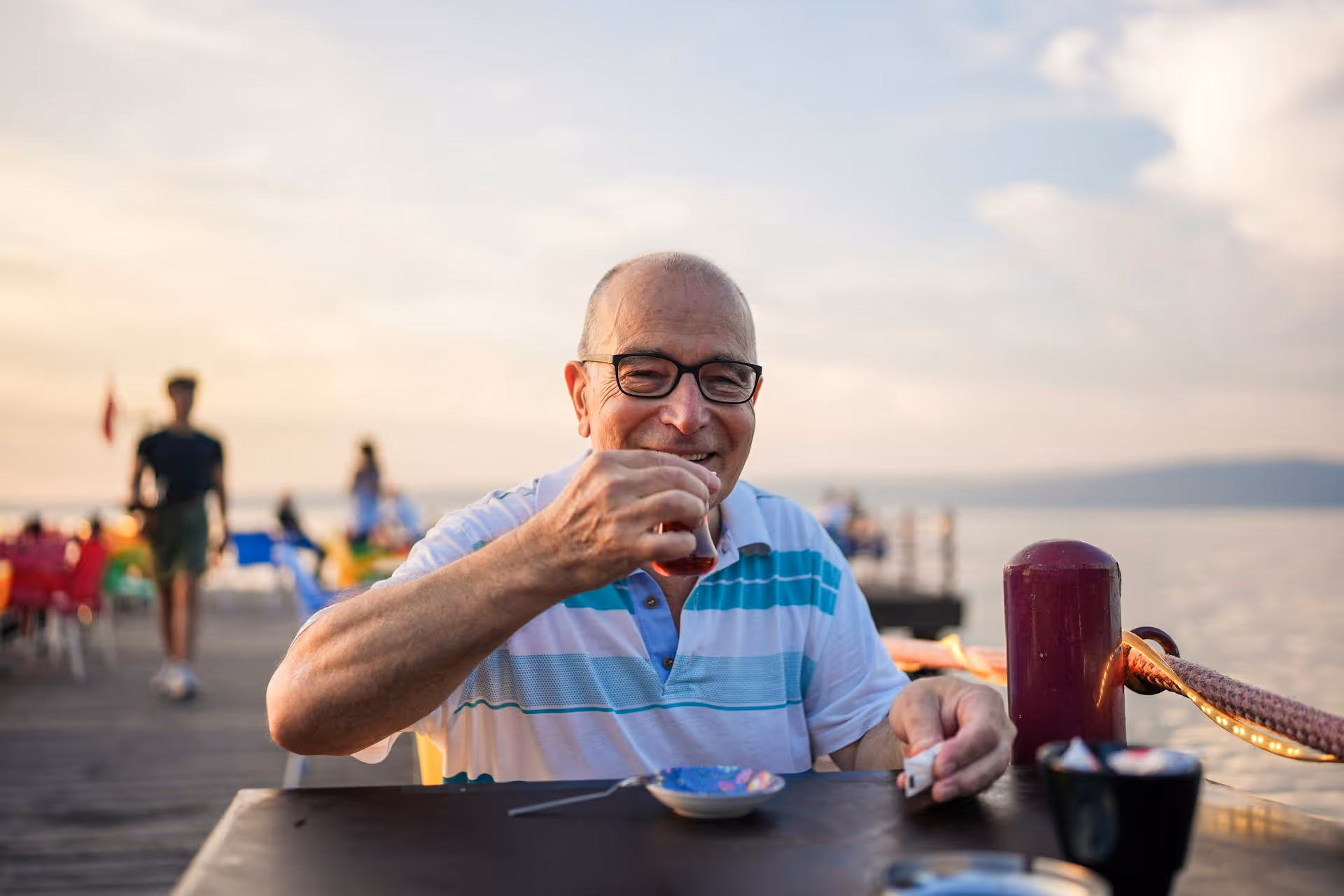Smiling elderly man wearing glasses and a striped polo shirt drinking tea at an outdoor table by the water during sunset.